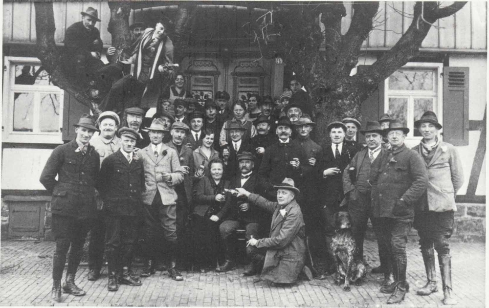 Männerchor Wahlscheid hilft beim Umzug von Fam. Breideneichen nach Auelerhof. Von links: 1. Max Fischer, Haus-Dorp; 2. Heinrich Breunsbach, Schuhmacher, Aggerhof; 3. Paul Fischer, Haus-Dorp und Hoffnungsthal 4. sitzend: Frau Hugo Breideneichen 5. sitzend: Hugo Breideneichen 6. knieend: Emil Weber, 1. Vorsitzender des Männerchores, Aggerhof 7. hinter 6. stehend: Gustav Hohn, Aggerhof 8. hinter dem Hund stehend: Hugo Fischer, Lebensmittel, Aggerhof 9. Hermann Schiffbauer, Gastwirt, Auelerhof 10. Gustav Schönenberg „Wieße“ Briefträger, Aggerhof 11. Paul Lemmer, Neuhonrath
2. Reihe: 1. mit Mütze heller Rock: Karl Weber, Auelerhof 2. mit Schlägermütze-dunkler Rock: Hermann Lindenberg, Müllerhof 3. ? 4. mit Mütze-eine Körperhälfte verdeckt: Wilhelm Steeger, Müllerhof 5. Friedchen?(Anna) Breideneichen 6. ? 7. Willi Hohn, Auelchen und Auelerhof 8. nur mit Oberkörper-heller Hut: Rudolf Kiel, Schiffarth 9. nur mit Kopf: Erwin Piel, Wahlscheid
hintere Reihen:schräg hinter der Dame mit Mütze u. weißem Hemd: Fritz Wilhelms (?), Auelerhof
vorletzte Reihe links – neben dem Kind – mit weißem Hemd: Karl Ober- dörster, Elektriker, Auelerhof
vorletzte Reihe Mitte – mit Zigarre im Mund: Karl Schiffbauer, Gastwirt, Auelerhof
vorletzte Reihe rechts – neben den beiden Damen – mit Hut: Karl Lindenberg, spät. Vorsitzender des Männerchores, Weeg neben dem Baum-vor den Kindern: Heinrich Naaf, Münchhof ganz hinten-mitten vor der Tür: Gustav Koser, Aggerhof
im Baum ganz rechts: Hugo Radermacher, Kreuznaaf; im Baum ganz links: Emst Frackenpohl, Weeg; im Baum Mitte: Rudolf Lindenberg, Münchhof
am Baum rechts-Kind: Emst Bergfelder, Müllerhof
